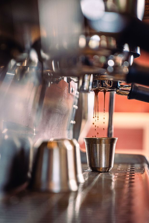 Close-up photograph of a commercial espresso machine dispensing coffee into a stainless-steel dosing cup. The image is tightly framed around polished metal equipment, portafilter, coffee droplets and the machine drip tray, with a warm, softly blurred café-like background. It is primarily a product/equipment detail rather than a view of a complete room.