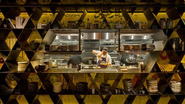 A dark, dramatic commercial restaurant interior viewed through a striking geometric shelving wall. The open kitchen is centered in the frame, with chefs working at a stainless-steel cooking counter, illuminated extraction canopies, and shelves displaying stacks of plates, bowls, cookware, bottles, and decorative objects. The shelving and surrounding surfaces feature bold triangular, diamond-like forms in black, brown, and gold tones.