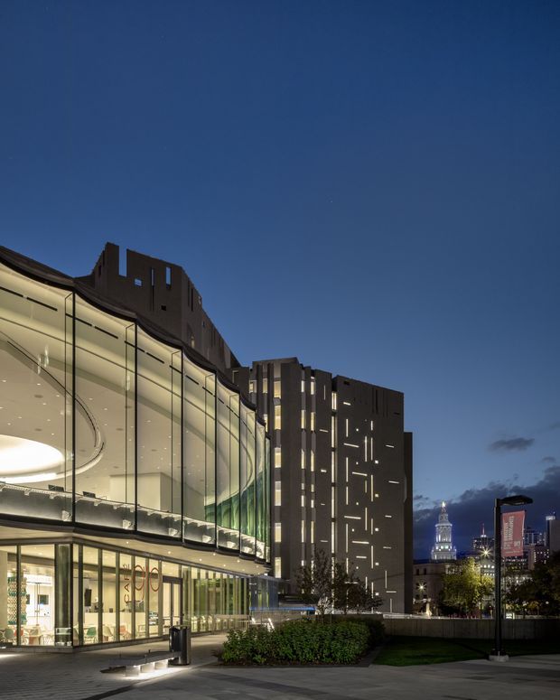 Dusk exterior view of a contemporary multi-storey commercial building in an urban setting. The curved glazed facade reveals a brightly lit interior, while a dark, rectilinear tower rises behind it with narrow vertical illuminated openings. Landscaped planting, paved pedestrian space, street lighting and a distant illuminated civic tower reinforce the city-centre context.