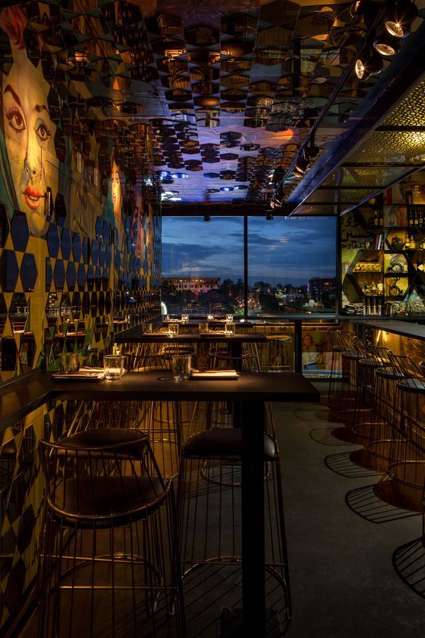 A dimly lit, highly eclectic bar or restaurant interior with a strong geometric and artistic design. Dark timber or black tables and slender metal-framed bar stools line the room, while the walls feature colourful geometric wall panels and large stylised portrait artwork. The mirrored or highly reflective ceiling creates repeated angular reflections of the room. A backlit bar with bottles and shelving occupies the right side, and large windows at the rear overlook an urban waterfront or cityscape at dusk. Warm pendant and track lighting produces a dramatic, moody atmosphere.
