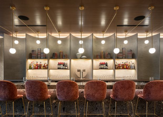 A sophisticated bar or hospitality interior viewed across a long stone bar counter, with a row of rust-coloured upholstered bar stools in the foreground. The space features mirrored or reflective dark shelving with displayed bottles and glassware, sculptural curved partition panels, warm timber ceiling lining, and multiple spherical pendant lights suspended from brass-toned fittings. The overall composition is symmetrical, refined, and warmly atmospheric.