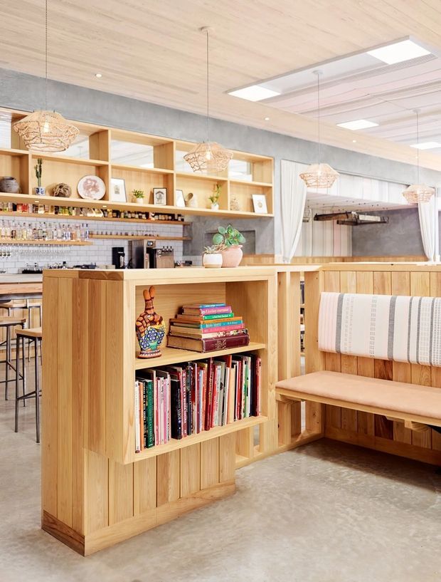 Bright contemporary hospitality-style interior featuring a timber-lined café or bar area with an open shelving display, books, bottles, a timber island partition, upholstered bench seating, pendant lights, concrete flooring, and exposed concrete wall elements. The space combines warm natural timber with light, airy finishes and integrated service counters.