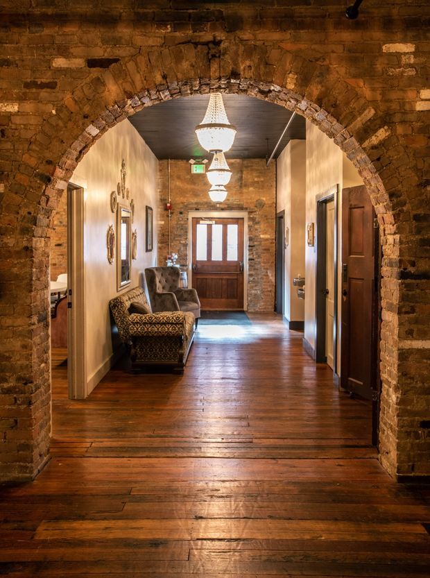 A warmly lit interior hallway viewed through a large exposed brick archway. The corridor has polished dark timber floorboards, exposed brick walls at the far end, painted cream walls, timber doors and trim, and a wood-panelled ceiling. Three hanging pendant chandeliers run along the hallway, with upholstered lounge chairs arranged along the left wall. A timber-framed glazed entrance door is visible at the end, creating a historic, character-rich atmosphere.