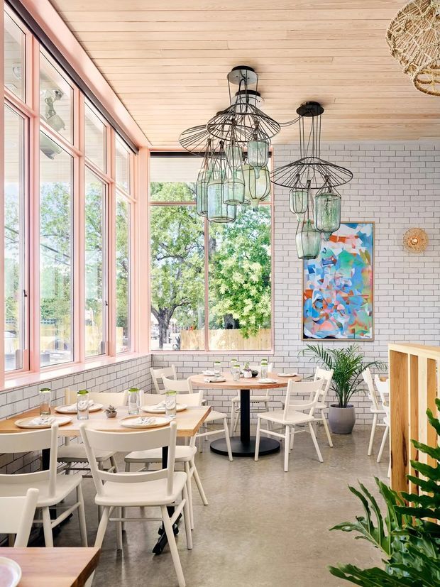 Bright contemporary dining area in a café or restaurant, featuring multiple timber dining tables with white chairs, large pink-painted framed windows, white rectangular tile walls, a light timber plank ceiling, polished concrete flooring, and distinctive suspended wire-and-glass pendant lights. Indoor plants and colourful artwork add decorative accents.