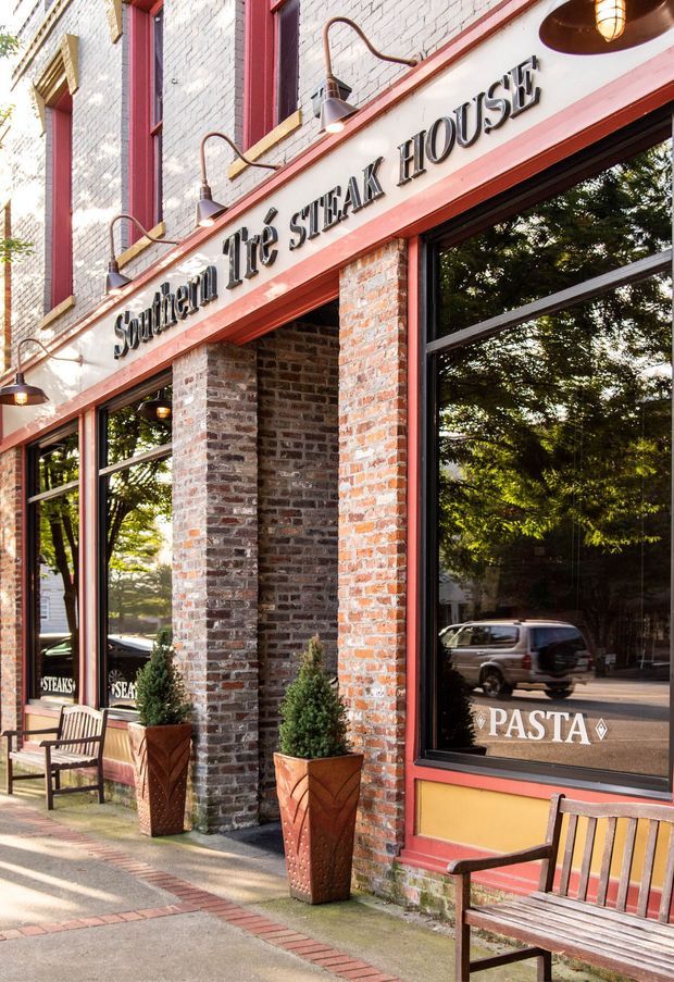 Street-level exterior view of a traditional restaurant storefront named Southern Life Steak House. The facade features exposed red brick, painted light brickwork, red-painted trim and window frames, large black-framed glazed windows, wall-mounted gooseneck lights, outdoor benches, and tall planters along the sidewalk. Restaurant signage and street reflections are visible.