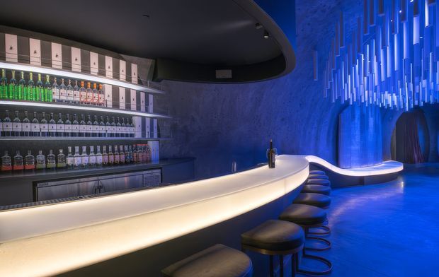 A dramatic, futuristic bar interior with a sweeping curved illuminated bar counter, rows of liquor bottles on open shelving, and a line of black bar stools. The space uses intense blue ambient lighting, textured dark walls, a sculptural ceiling, and a hanging installation of vertical illuminated elements, creating a nightclub-like atmosphere.