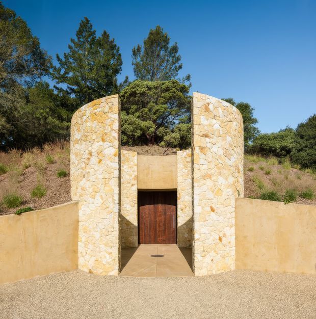 A sculptural exterior entrance set into a landscaped earth mound. Two tall, curved walls clad in irregular pale natural stone frame a recessed timber double door, with smooth rendered retaining walls extending outward. Native trees and grasses surround the structure beneath a clear blue sky.