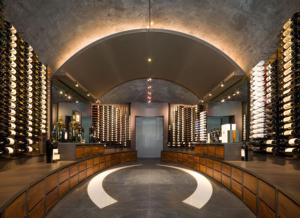 A dramatic, symmetrical wine cellar or wine retail display room with extensive bottle racks lining both side walls and rear shelving. Curved vaulted ceilings, warm concealed cove lighting, dark stone or concrete-look finishes, timber-toned cabinetry, glass-fronted display areas, and a circular contrasting floor inlay create a luxurious, immersive atmosphere.