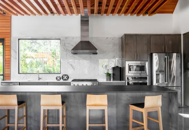 A contemporary kitchen viewed straight on, featuring a large dark island with a thick countertop and integrated seating for four. The kitchen has dark wood cabinetry, stainless-steel appliances, a marble-look tiled splashback, a broad window over the sink, and a striking exposed timber batten ceiling with visible recessed lighting. Light timber bar stools add contrast to the dark cabinetry and island.