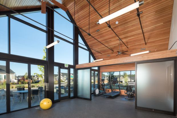 A bright, contemporary communal fitness room with floor-to-ceiling black-framed glazing, a dramatic sloped timber-lined ceiling, exposed dark structural beams, and suspended linear lighting. Exercise equipment is arranged near the glazed wall, with an outdoor patio and residential development visible beyond the windows.