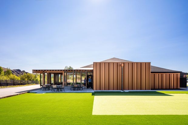 Single-storey contemporary building viewed from the landscaped exterior, with a distinctive ribbed copper-brown metal-clad central volume, dark roof, extensive floor-to-ceiling glazing, and a covered outdoor dining terrace. A broad bright green lawn or synthetic turf dominates the foreground, with low fencing and a dry natural landscape beyond.