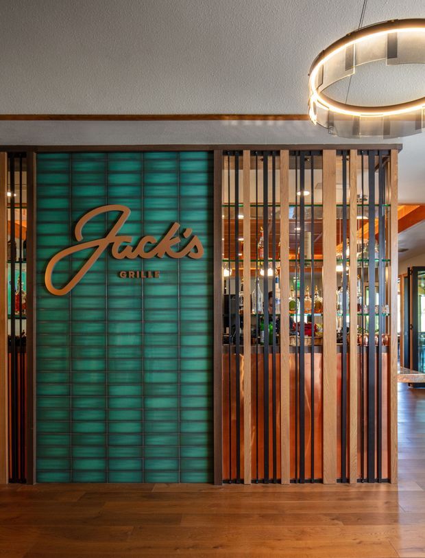 Interior view of a restaurant branded “Jack’s Grille,” featuring a prominent illuminated turquoise tiled feature wall with raised lettering, flanked by a vertical timber and dark metal slat partition. Warm timber flooring, wood trim, a circular pendant light, and glimpses of the dining/bar area create a polished hospitality interior.