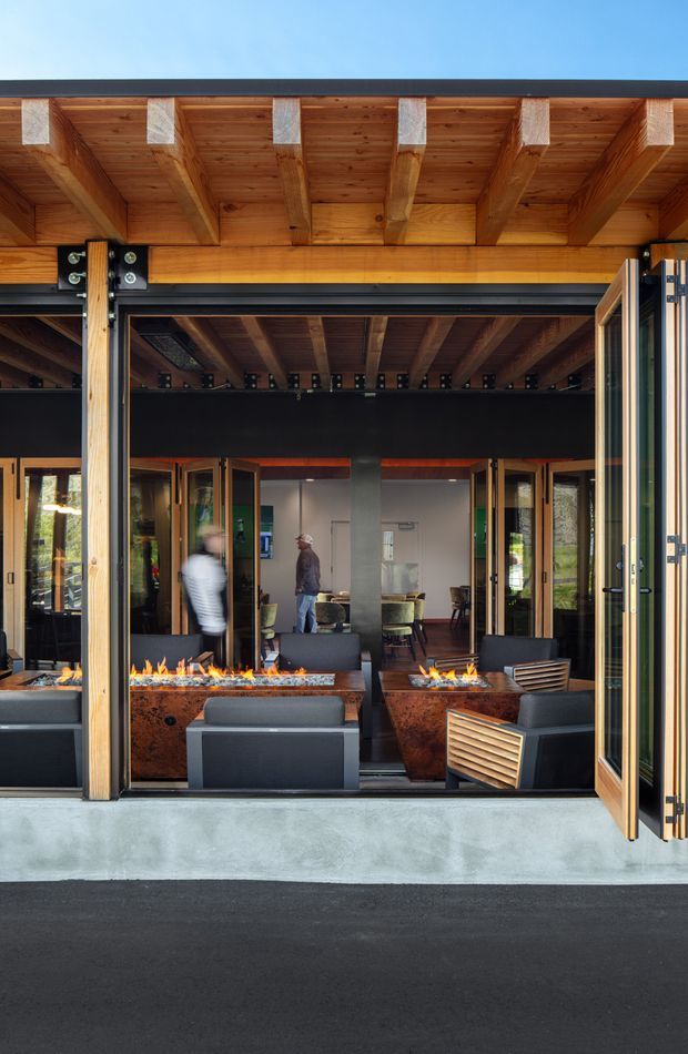 View from outside into a contemporary hospitality-style interior through fully opened timber-framed glazed doors. The building features a deep exposed timber beam and soffit canopy, dark steel structural framing, concrete threshold, and large indoor-outdoor opening. Inside are dark lounge chairs, rusted metal rectangular fire features with visible flames, dining tables, and exposed structural beams.
