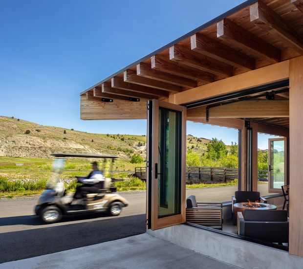 A contemporary open-sided architectural space with a deep timber-framed roof overhang, exposed timber rafters, large folding or sliding glazed doors, and a polished concrete floor. The space opens onto a paved road or driveway and a landscaped hillside setting. Outdoor lounge furniture and a fire feature are visible inside, while a golf cart passes outside. The image highlights the timber structure, indoor-outdoor connection, and expansive operable glazing.