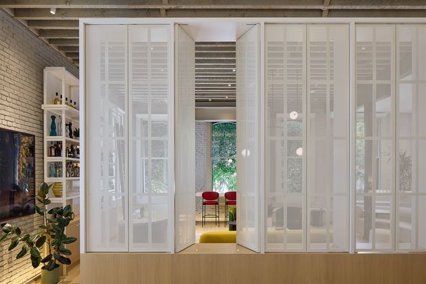 Interior view of a bright contemporary living area screened by full-height white translucent framed partitions with louvred or shutter-like panels. A central opening leads to a compact dining or sitting area with red chairs and a yellow-green upholstered bench. The space has exposed timber ceiling beams, pale brick walls, light timber cabinetry or a low partition, and abundant greenery visible through glazing. A television, shelving, and indoor plants occupy the left side.