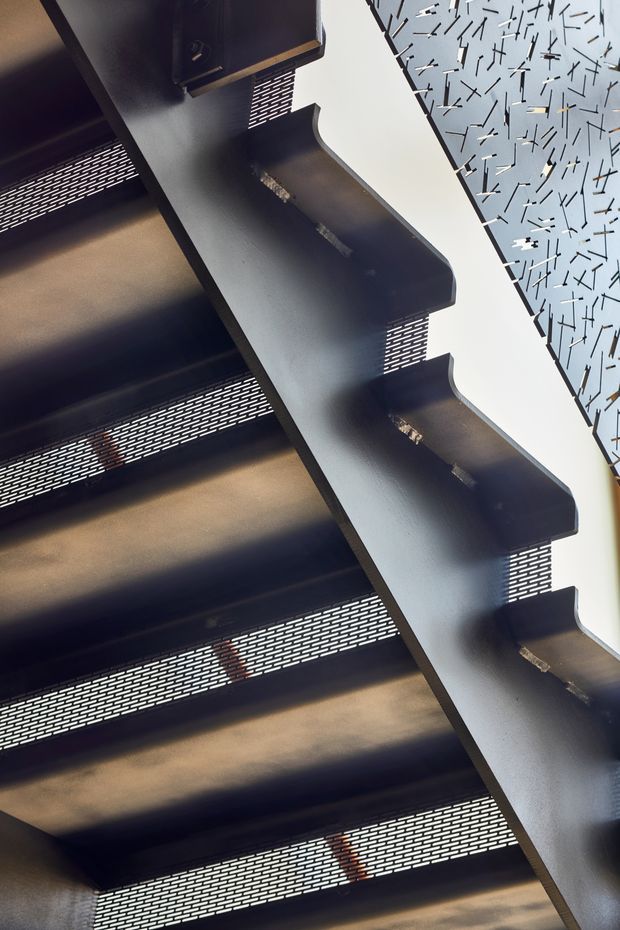 Close-up upward-looking architectural view of a dark metal stairway with perforated or expanded-metal treads and a heavy zigzag-edged side stringer. A pale patterned wall or screen with scattered linear marks is visible alongside the stairs. The image is dominated by industrial metal construction and strong diagonal geometry, with no furniture or room context visible.