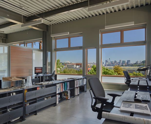 A contemporary open-plan office with exposed corrugated metal ceiling decking, dark structural beams, polished concrete flooring, large timber-framed windows, and extensive built-in shelving and workstations. The space has panoramic urban views and a clean, industrial studio atmosphere.