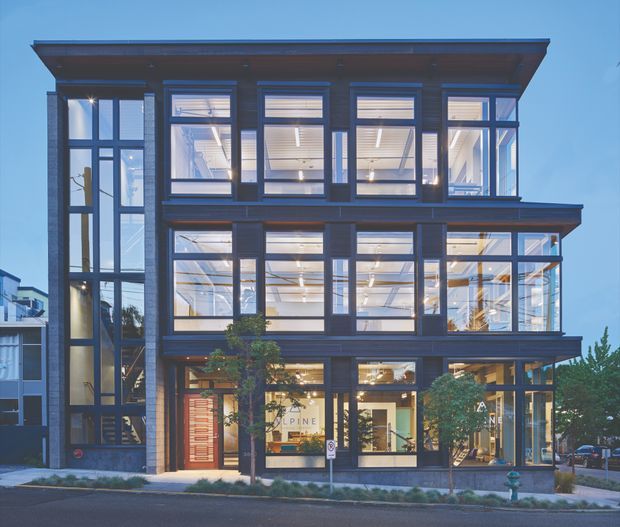 Street-facing view of a contemporary multi-storey commercial or mixed-use building with a dark framed facade, extensive floor-to-ceiling glazing, projecting horizontal roof and canopy lines, and a tall glazed stairwell or circulation volume at the left. Warmly illuminated interior spaces and ground-floor retail or office glazing are visible.