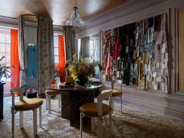 Eclectic, richly layered dining room with a round dark stone or marble dining table, upholstered vintage-style chairs, patterned curtains, large windows and doors, and a dramatic wall-mounted textile or fabric sample display. The room features ornate moulded wall and ceiling trim, a reflective metallic-toned ceiling, pendant lighting, and a plush neutral carpet. The overall atmosphere is colourful, artistic, and highly decorated.