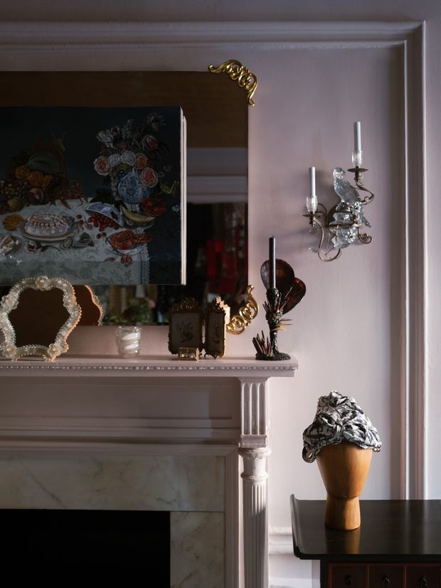 A close-up of a classically detailed fireplace mantel and mirror in a softly lit interior. The pale painted mantel features fluted pilasters, moulded trim and a marble fire surround, while an ornate gold-framed mirror, decorative silver wall sconce, sculptural objects and a dark side table create a refined, eclectic composition.