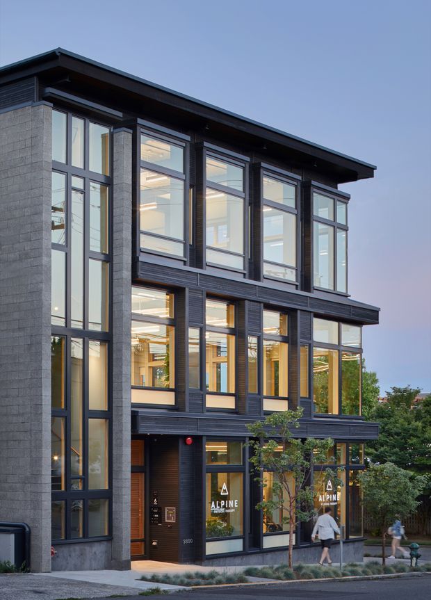 Exterior photograph of a contemporary multi-storey commercial building with a strong vertical facade rhythm. The building combines dark metal or timber-look cladding, large floor-to-ceiling glazed windows, and grey masonry side walls beneath broad flat roof overhangs. Ground-floor storefront glazing and visible signage indicate a commercial or office tenancy, with a landscaped pedestrian frontage.