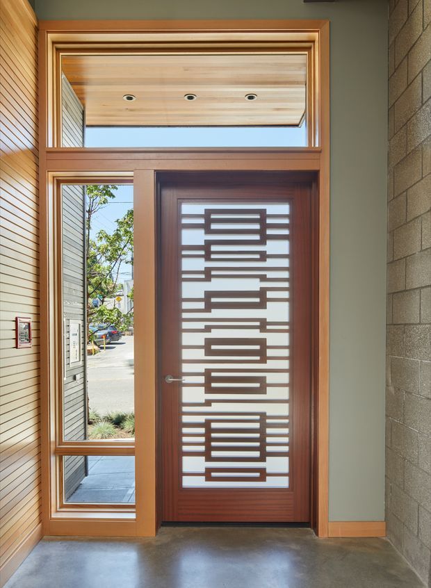 Modern entryway featuring a tall timber-framed glazed door with a geometric timber grille and frosted or translucent glazing. Matching timber sidelights and a transom bring in daylight, while the entry has a smooth concrete floor, timber-lined wall and ceiling surfaces, and an exposed grey concrete-block wall.