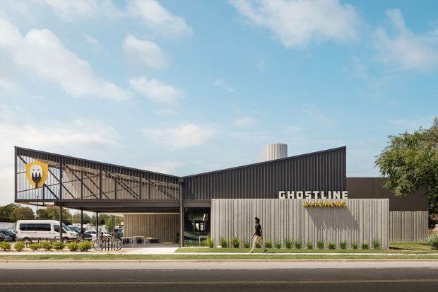 Exterior street view of a contemporary commercial building with a dark corrugated metal facade, a large elevated steel-framed sign and graphic panel, and a lighter vertically ribbed entry volume. The building has an open covered forecourt with bicycles and parked vehicles nearby, landscaped planting along the frontage, and prominent signage reading “GHOSTLINE KITCHENS.”