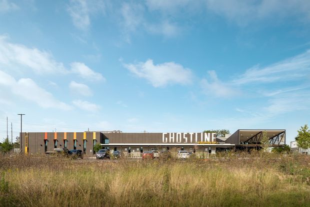 Wide exterior view of a low-rise commercial building set behind a field of tall grasses. The building has a long, dark brown facade with colorful vertical accents, large white “GHOSTLINE” signage, and a prominent open steel-framed upper structure at the right. Utility poles and a bright blue sky form the background.