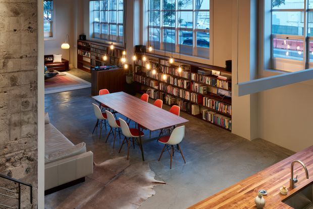 A spacious open-plan interior with a dining and reading area. A long timber dining table with colourful molded chairs sits on a polished concrete floor, beside extensive built-in bookshelves illuminated by pendant bulbs. Large industrial-style windows bring in daylight, while a partial kitchen benchtop with sink is visible in the foreground. A lounge area and bedroom-like nook appear deeper in the space. The interior combines exposed concrete, timber, metal-framed glazing and warm ambient lighting.