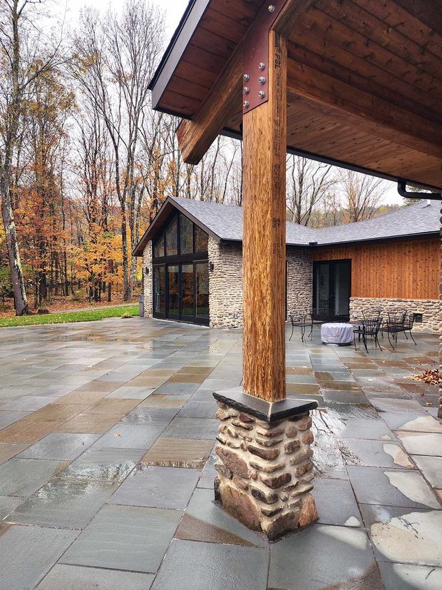 A rustic covered outdoor patio beside a low-slung house in a wooded setting. The structure features exposed timber posts and a timber-lined roof soffit, stone-clad column bases and walls, large dark-framed glazed doors, warm timber wall cladding, and a broad paved terrace with outdoor dining furniture. The architecture has a strong timber-and-stone lodge character and appears to be photographed after rain.