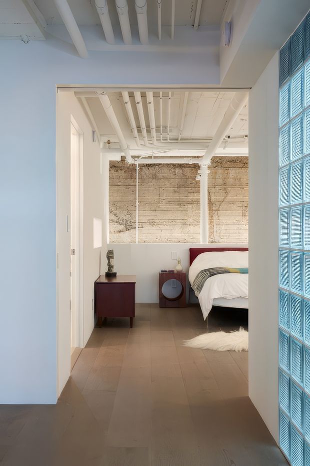 View through a wide doorway into a bright bedroom with an exposed industrial-style ceiling of painted white pipes and beams. The room has a partial-height wall, textured raw concrete or plaster feature wall, warm neutral flooring, a deep red upholstered bedhead, dark timber bedside furniture, and a blue glass-block or louvred wall at the right edge. The space combines utilitarian architectural elements with refined contemporary furnishings.