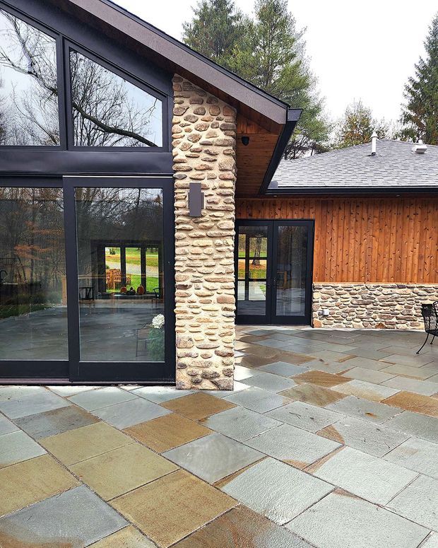 Exterior view of a contemporary timber-and-stone house opening onto a broad paved terrace. The building features dark-framed floor-to-ceiling glazing, a prominent rounded natural-stone structural pier, vertical timber wall cladding, exposed timber soffits, and a low-pitched shingle roof. Large rectangular stone pavers form the outdoor patio, with interior spaces visible through the glazing.