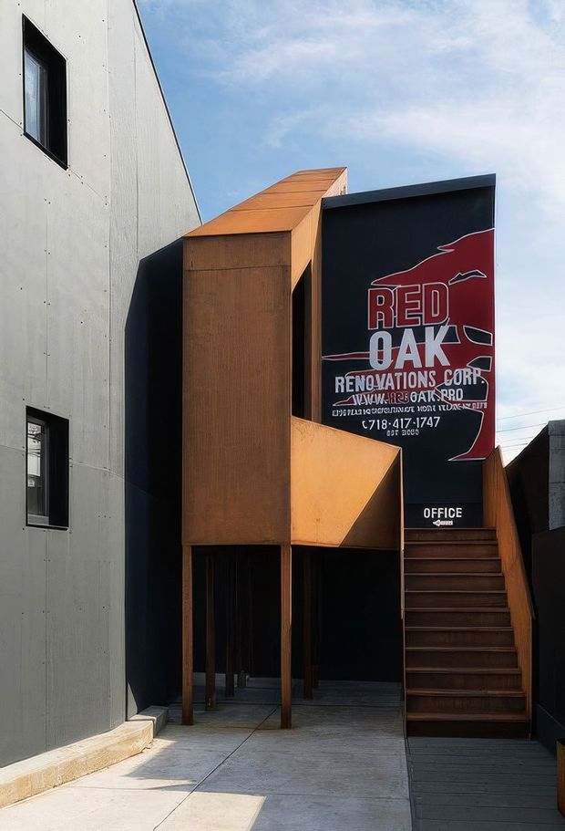 A compact contemporary commercial office facade viewed from an exterior walkway. The building combines a pale grey panel-clad wall with bold black geometric volumes, warm rust-coloured metal or timber-like cladding, a projecting upper form, and an external timber-toned staircase leading to an office entrance. A large red-and-white Red Oak Renovations Corp sign dominates the upper facade. The composition has angular, sculptural forms and strong contrasting materials under bright daylight.