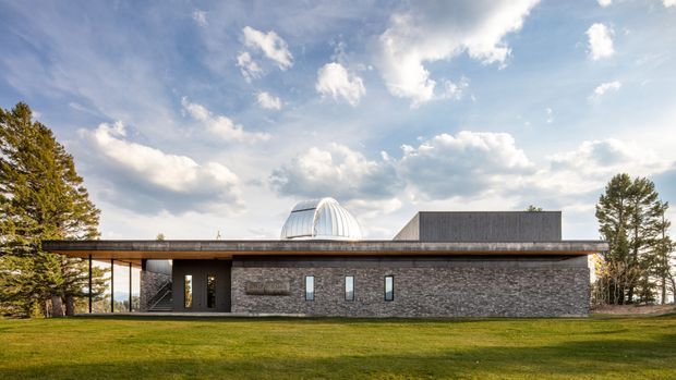 A low, single-storey modern building set in a broad landscaped lawn, with a long dark stone-clad facade, a thin projecting flat roof, narrow vertical windows, and a covered open-sided entrance or walkway. A distinctive ribbed domed roof element rises behind the main roofline, while mature trees and an expansive sky surround the building.