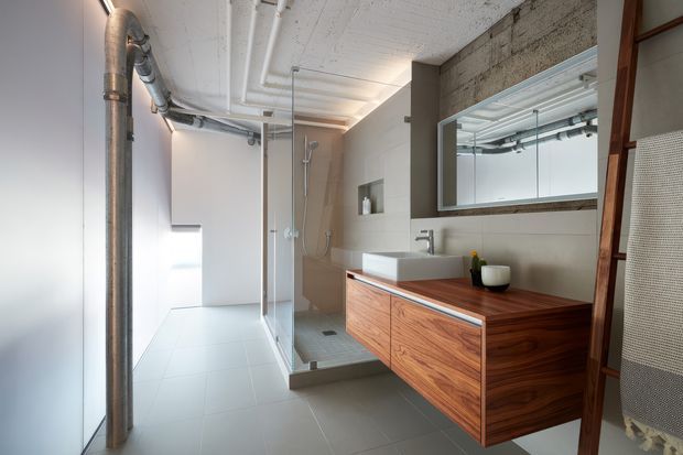 Modern industrial-style bathroom with a walk-in glass shower, wall-mounted timber vanity, large rectangular mirror, exposed metal plumbing, and a raw concrete ceiling and feature wall. The neutral grey and warm wood palette creates a clean, refined atmosphere.