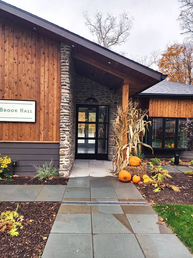 Exterior view of a low, modern building entrance with warm vertical timber cladding, dark trim, a broad roof overhang, natural rough-stone wall sections, and black-framed glazed doors and windows. A paved path leads through landscaped garden beds decorated with pumpkins and dried corn stalks. A sign reading “Brook Hall” is mounted on the facade.