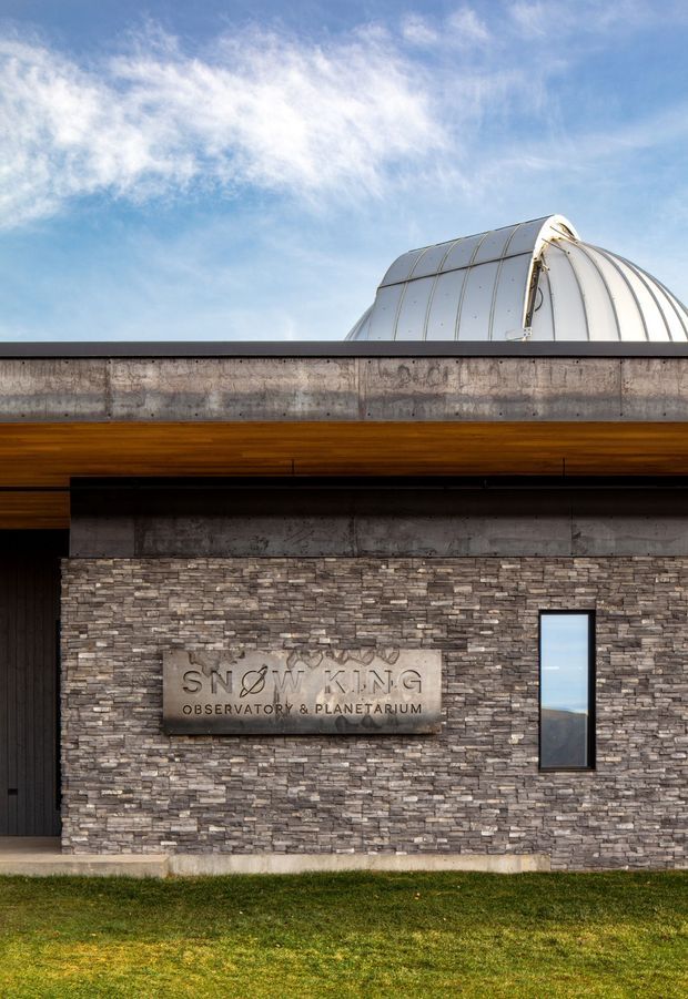 Exterior architectural photograph of the Snow King Observatory & Planetarium. The building features a horizontally layered facade with grey stacked-stone cladding, dark metal trim, warm timber soffits, narrow vertical glazing, and a prominent silver domed observatory roof visible above the parapet. A mounted metal sign identifies the facility, with lawn and blue sky surrounding the structure.