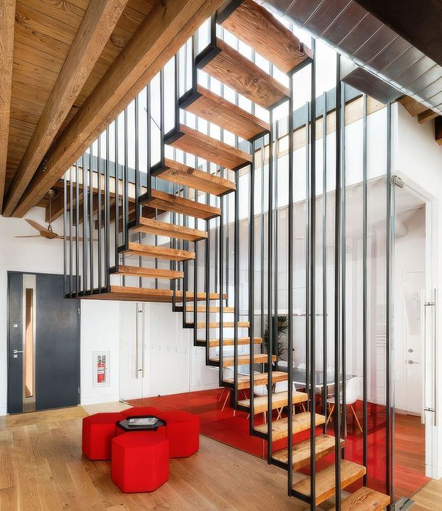A bright, contemporary interior centered on a sculptural open-riser staircase with warm timber treads and substantial timber ceiling beams, enclosed by slender black metal framing and vertical glazing. The staircase rises through a double-height space, with a vivid red carpeted or painted floor zone and matching red upholstered ottomans around a small black table. White walls, black doors, exposed timber structure, and abundant glazing create a dramatic loft-like composition.