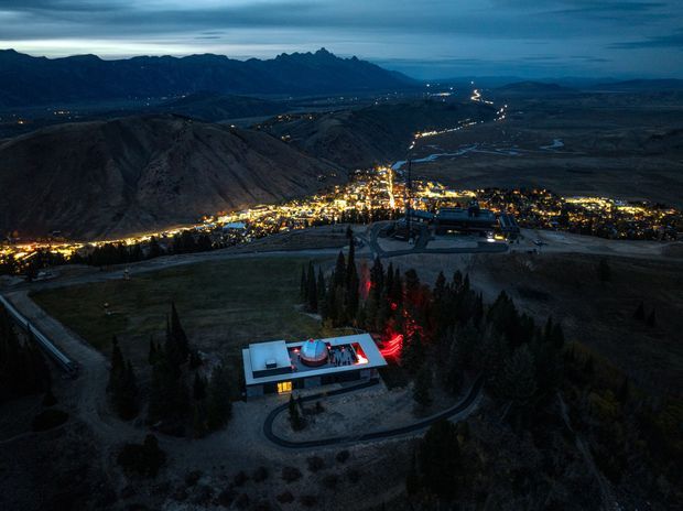 A dusk aerial view of a modern isolated building on a wooded mountainside, with illuminated outdoor areas and a winding access road. A broad valley town and meandering lights extend below, backed by rugged mountain ranges under a dark blue evening sky.