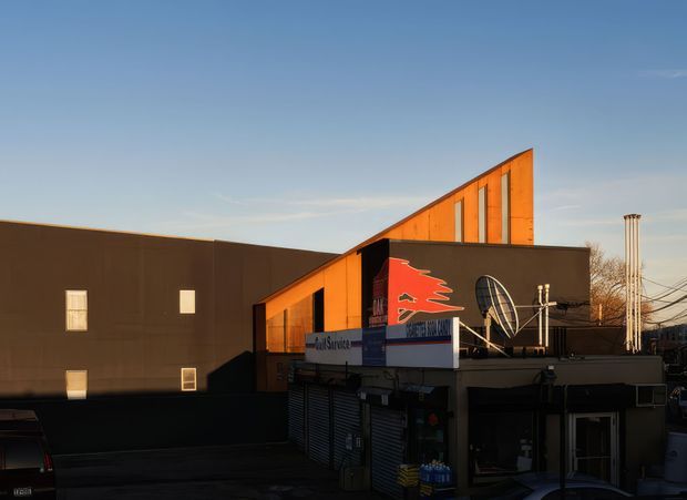 Exterior view of a compact urban commercial building in low evening light. The facade combines dark brown wall panels with a prominent angular orange upper volume and narrow vertical windows. Ground-level shopfronts, signage, security shutters, rooftop equipment, utility wiring, and a satellite dish are visible. The composition emphasises the building facade and its strongly geometric roofline against a clear blue sky.