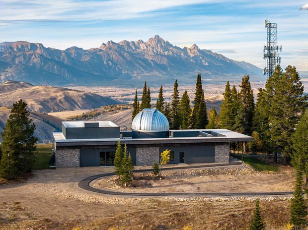 A low-slung contemporary building set on a mountainous, elevated site, surrounded by coniferous trees and expansive rugged alpine terrain. The dark masonry and metal-clad facade has broad horizontal glazing, a flat roof, and a prominent silver domed roof element at the centre. A communications tower stands beside the building, with a curved driveway in the foreground.