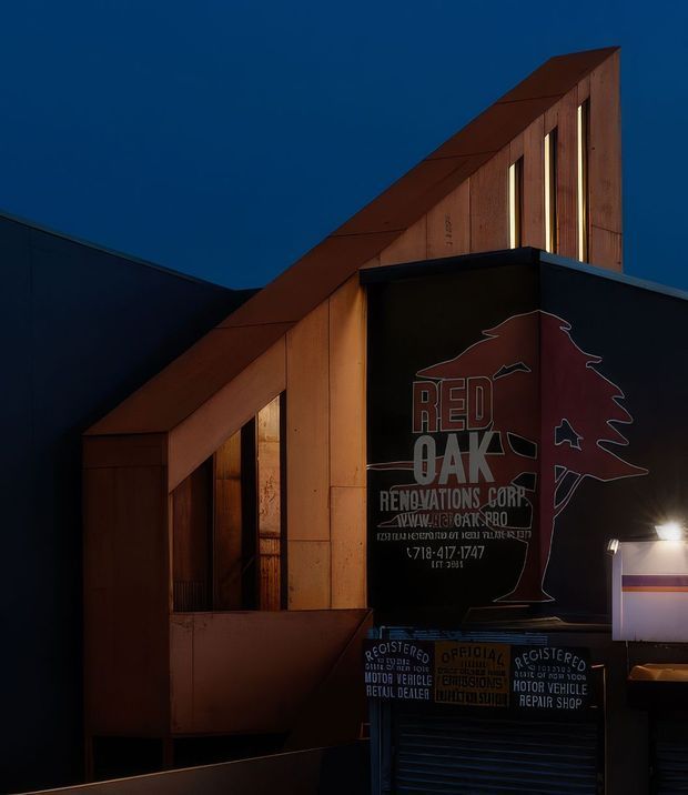 Night-time exterior view of a small angular commercial building with a distinctive sloping roofline and warm timber-clad facade. Dark painted wall surfaces contrast with the illuminated timber panels and narrow vertical windows. A prominent sign identifies the premises as Red Oak Renovations Corp, with additional automotive-related signage near the entrance.