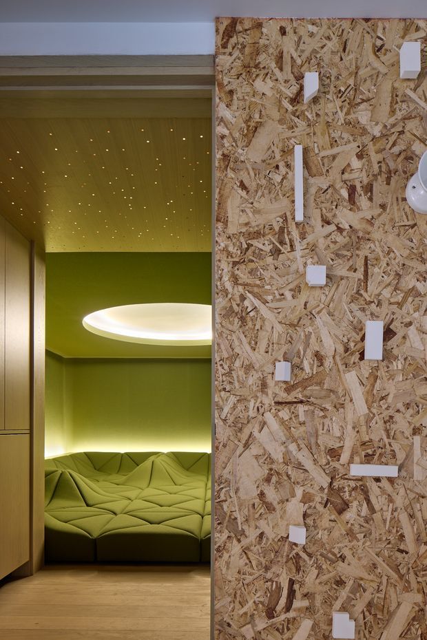 A vertically framed contemporary interior detail showing a small green upholstered seating nook behind a partially open timber-panelled door or partition. The nook has a sculptural tufted sofa, a green wall and ceiling with numerous tiny recessed lights, and a large glowing oval pendant light. The foreground wall is clad in exposed oriented strand board (OSB) with scattered white rectangular fixtures or controls. Timber flooring and warm concealed lighting create a dramatic, playful atmosphere.