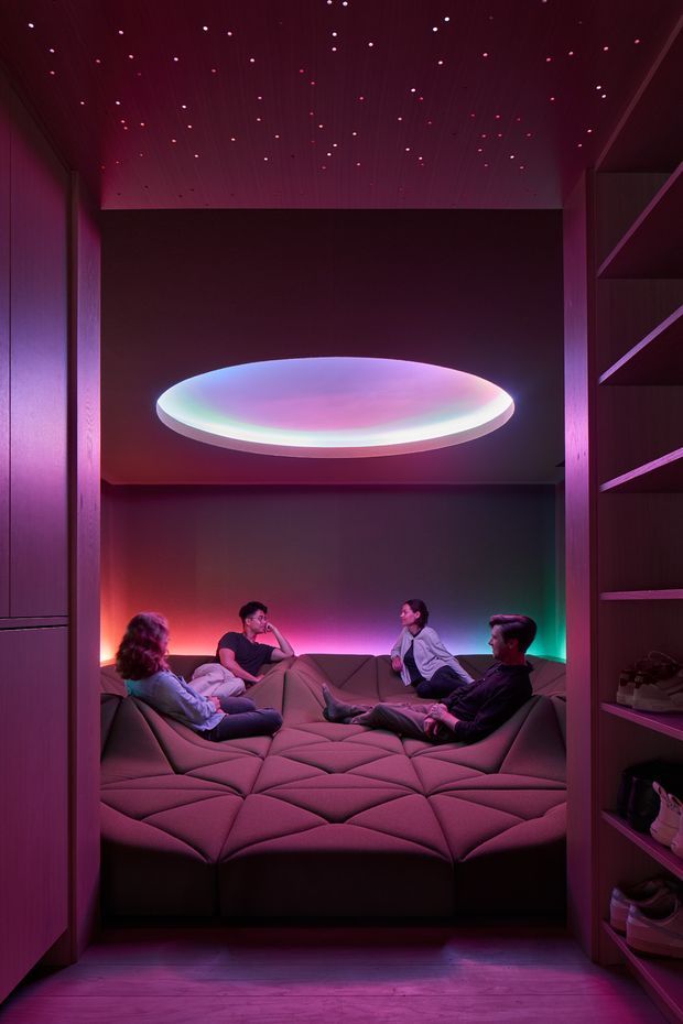 A compact, enclosed media lounge with a large modular padded sectional platform surrounded by built-in shelving. The space features dark timber wall and ceiling finishes, vivid magenta, red, blue, and green ambient lighting, a glowing oval ceiling light, and a star-like pinpoint light installation across the ceiling. Several people are seated informally on the lounge, creating an immersive, futuristic home-cinema atmosphere.