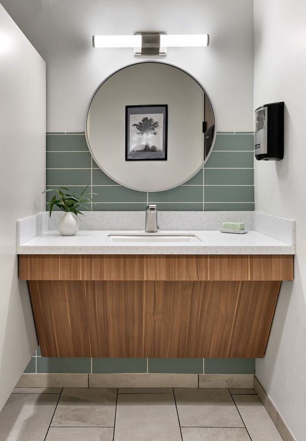 A compact contemporary bathroom vanity viewed head-on. The room features a floating, warm wood-grain vanity with a white speckled countertop, integrated rectangular basin, chrome faucet, large round mirror, and horizontal green-grey tiled splashback. White painted walls, pale floor tiles, a linear wall light, soap dispenser, and small plant complete the clean, modern scheme.