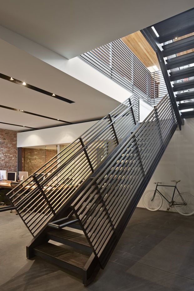 Interior view of a dramatic industrial-style stairway with dark metal open-tread stairs and dense horizontal metal balustrades. The stair rises through a tall void beneath a slatted skylight or clerestory, with exposed warm brick walls, dark tiled flooring, linear recessed lighting, timber ceiling lining, and a bicycle displayed beside the stairs. Office workstations are visible in the background.