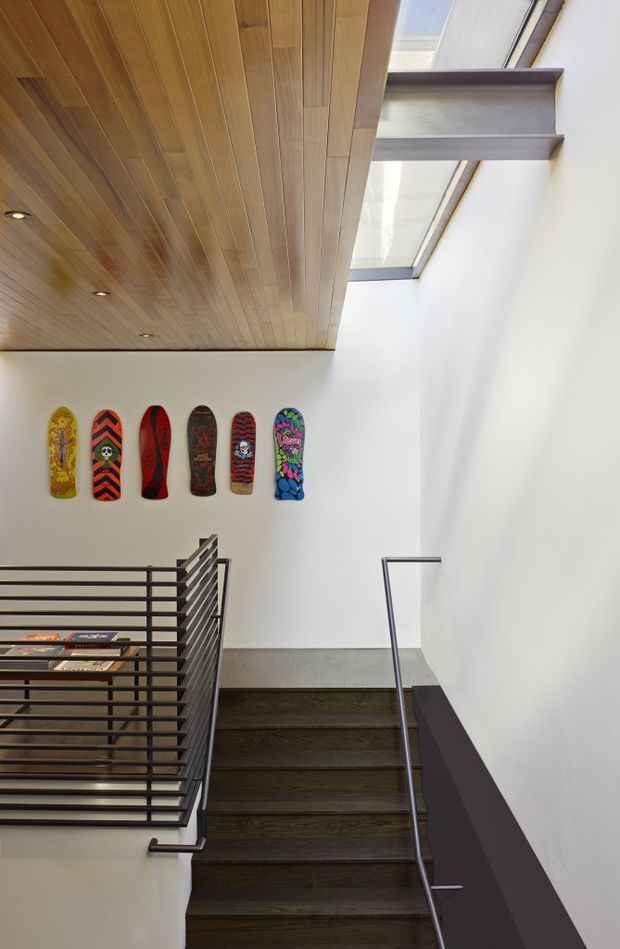 Interior view of a modern stairway rising to a landing, with dark timber stair treads, slim metal handrails, and a horizontal metal balustrade. A white double-height wall is decorated with colourful skateboard decks, while a warm timber-panelled ceiling with recessed spotlights extends overhead. Clerestory-style roof glazing brings in bright natural light.