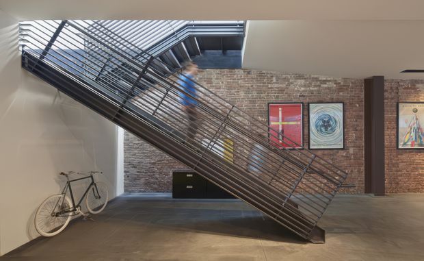 Interior view of a contemporary industrial-style entry or circulation space dominated by a dramatic steel staircase with open metal balustrades and a skylight above. Exposed red brick walls, polished concrete flooring, dark structural columns, framed artwork, and a bicycle create a loft-like atmosphere.
