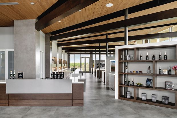 A spacious contemporary commercial interior, likely a tasting room or hospitality venue, with a long reception or service counter in the foreground, open shelving displaying bottles and glassware, exposed dark timber beams, a warm timber-lined ceiling, large glazed openings, and a polished concrete floor. The space has a refined industrial character with strong indoor-outdoor visual connection.