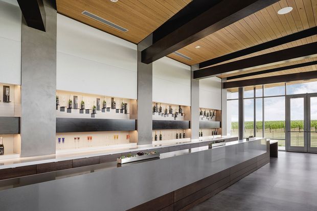 A spacious contemporary tasting room or commercial hospitality space with a long dark island counter, built-in display shelving for bottles and glassware, pale wall panels, exposed dark structural beams, and a warm timber slatted ceiling. Full-height glazed doors and windows open to a landscaped vineyard-like setting. The interior has a refined, minimalist character with strong linear geometry and abundant natural light.
